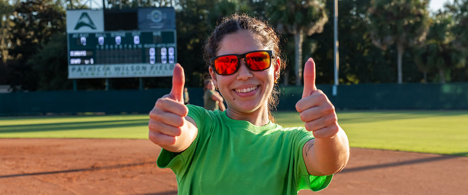 student with sunglasses giving two thumbs up on the softball field