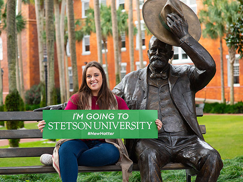 student with sign that says going to stetson sitting next to statue of John B. Stetson
