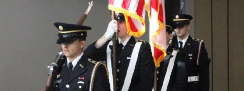 Three individuals in military uniforms maching with flag