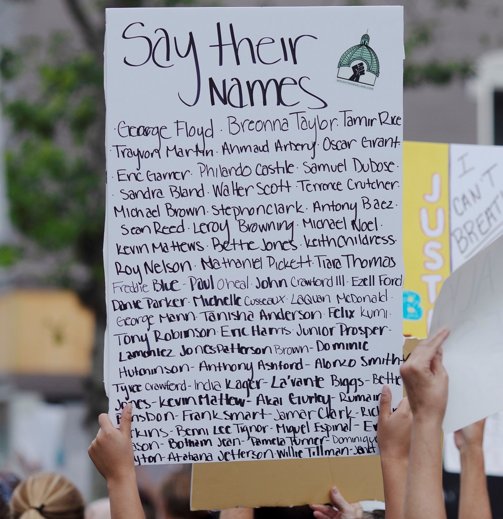 BLM protester holding a sign with the header 'Say Their Names', followed by a list of individuals, advocating for the recognition and remembrance of their lives.