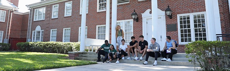 8 students sitting on the steps of hollis hall, a red brick building with white columns