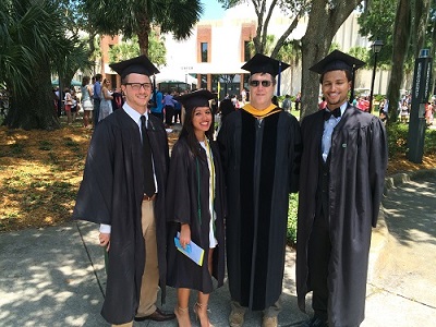 A group three smiling students along with a faculty member in a black robe and a gold stole