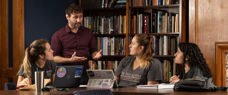 a professor gestures while speaking to three focused students in a library