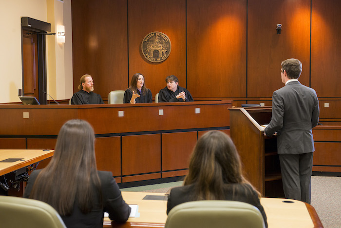 Two female students watch as a male student stands at podium addressing Professor Long, Bowman, and Vaughan