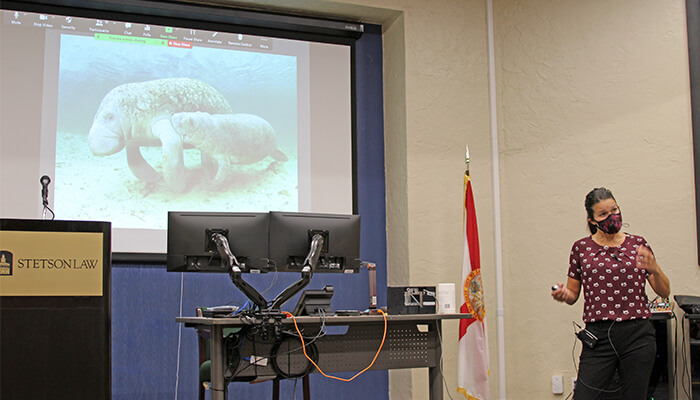 The speaker stands in front of a projector screen with manatees