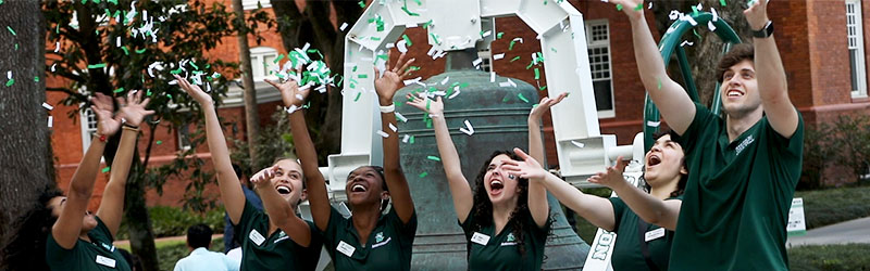 Group of enthusiastic students in green shirts tossing confetti in the air on campus, celebrating an event with joy and excitement.