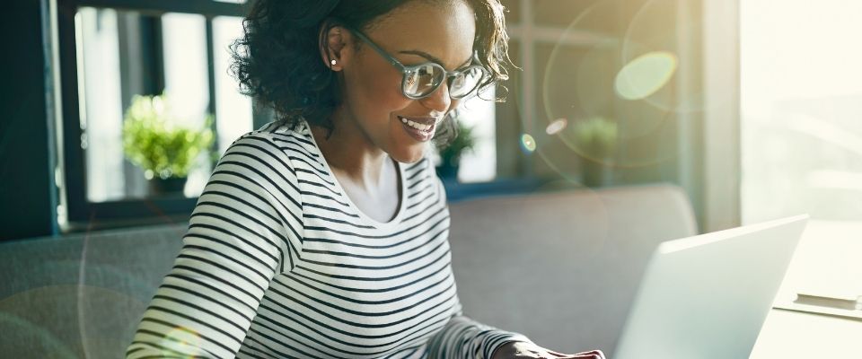 A young women sitting in her home smiling while she works on her laptop.