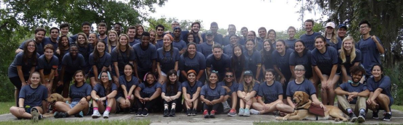 Large group of student in matching shirts posing outdoors on a path under leafy trees with two dogs.