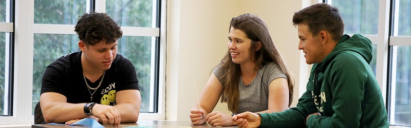 Three students sit around a table in a bright classroom, smiling and talking in group discussion.