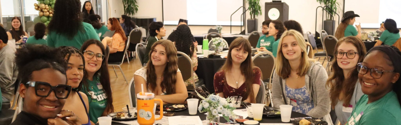 Students sitting around a circular table, enjoying a meal while facing a camera