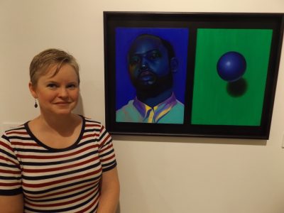 Hand Art Center director Tonya Curran poses beside “Self Portrait in Blue” by Trent Tomengo,” one of the works in the exhibit “Alumni Permanent Collection Highlights.”