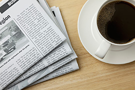 A cup of black coffee sits beside a newspaper on a table.