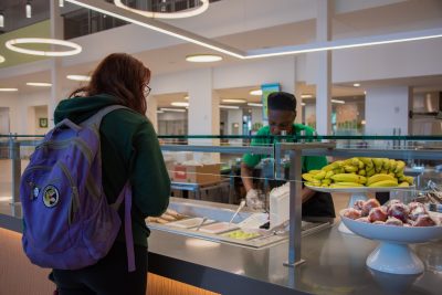 A dining worker dishes food into a to-go container for a waiting student