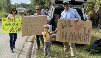 Adults and a child are holding signs, We Love you Hannah, Welcome Home
