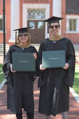 mother and son pose in cap and gown, holding diplomas, on campus