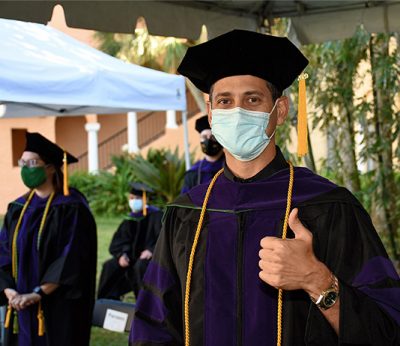 A law graduate gives a thumbs up in cap and gown, and face covering