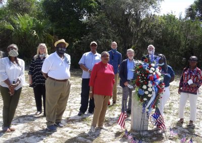 A group of people stand around a grave marker adorned with flags and red and white flowers.